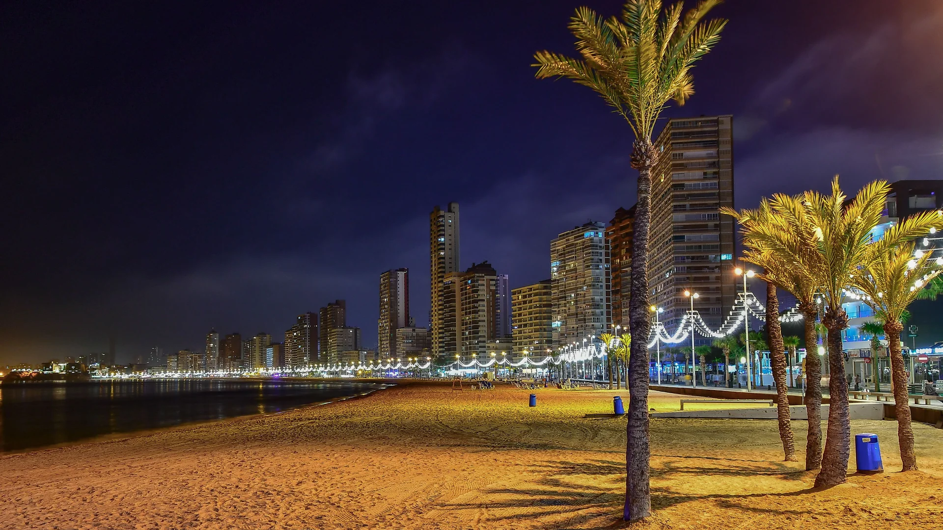 Palm trees and strings of lights along Benidorm's beach at night, with the lit high-rise skyline behind them